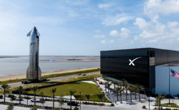 a large silver spacecraft rolls down a seaside road next to a large black building