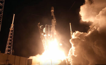 a white and black rocket launches into the night, its orange and white engine plume lighting the ground below