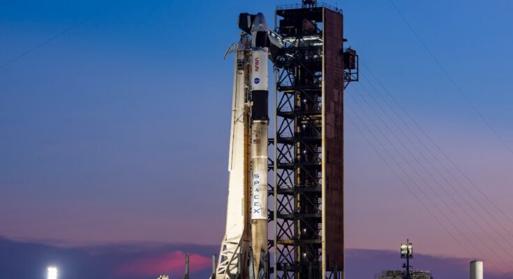 a white rocket stands upright on a launch pad at sunset