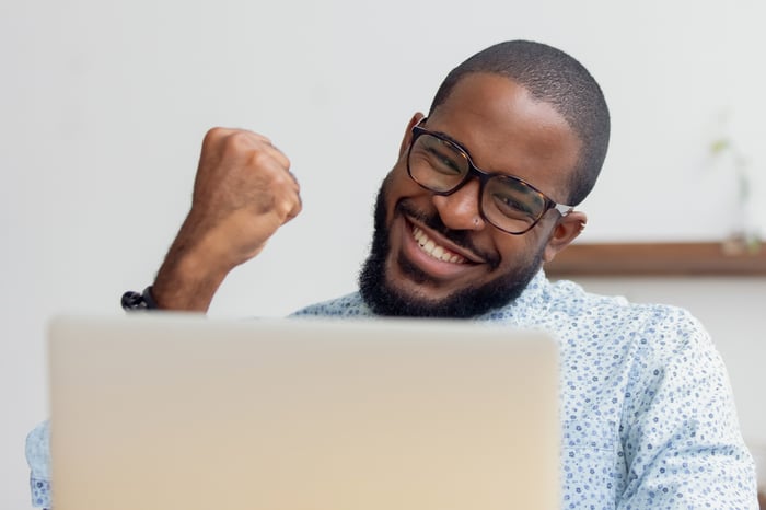 Una persona sonriente sosteniendo un puño mientras mira una computadora portátil.