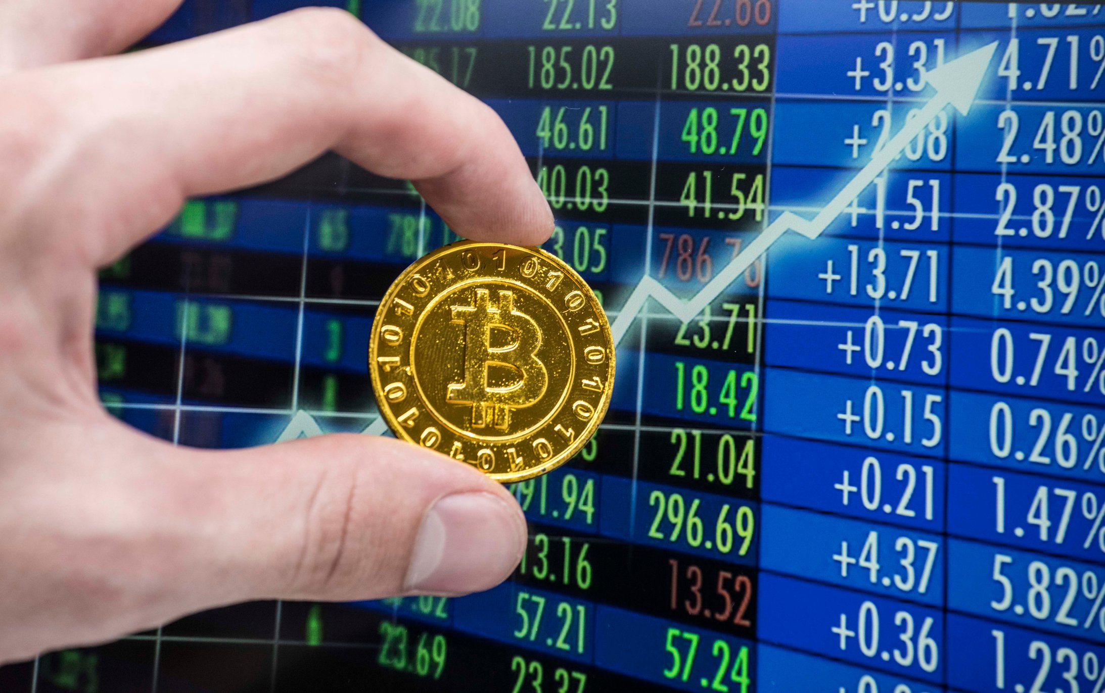 Una mano que sostiene un Bitcoin dorado junto a una flecha hacia arriba en una pantalla de operaciones.