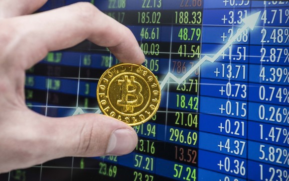 Una mano sostiene una moneda de oro con el logotipo de Bitcoin en una pantalla con un gráfico de acciones en aumento y otros datos sobre el precio de las acciones.