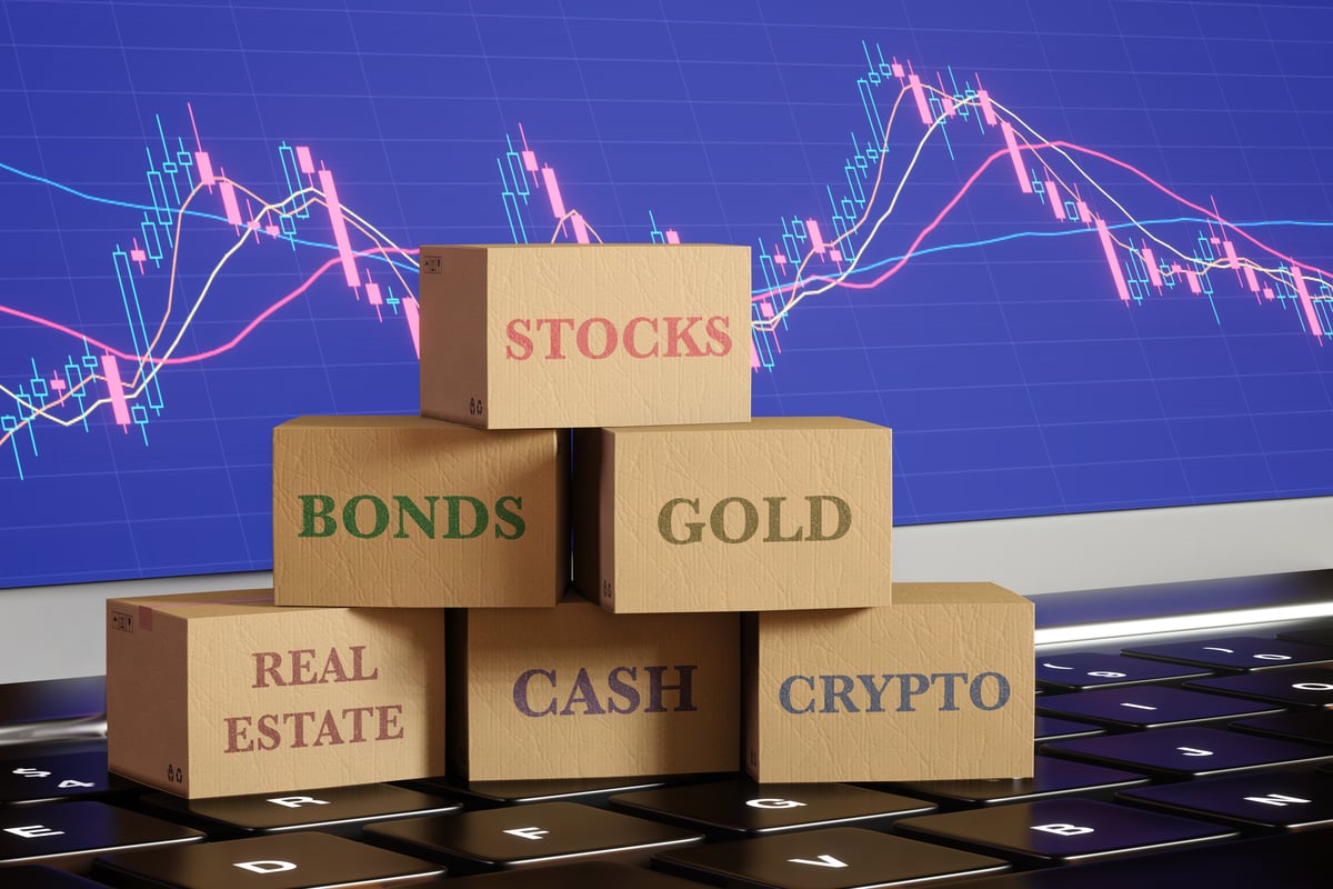 Una pila de bloques de madera sobre un teclado que muestra clases de activos que incluyen acciones, bonos, oro, bienes raíces, efectivo y criptomonedas, con un gráfico de velas al fondo. 