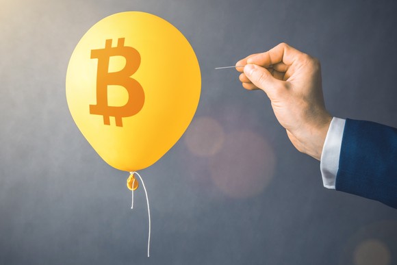 Un globo con el logo de Bitcoin flota junto a una mano que se acerca sosteniendo un alfiler.