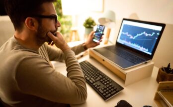 Person looking at a chart on their computer screen while holding a smartphone.
