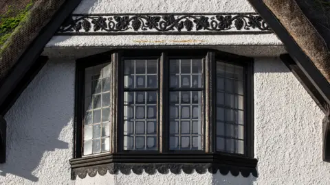 Archivos históricos de Inglaterra Una vista ampliada de una de las ventanas de un edificio con techo de paja. El marco de la ventana está pintado de marrón y hay un patrón encima de la ventana. 