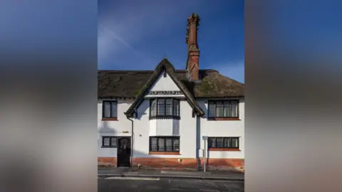 Archivos históricos de Inglaterra Parte del gran edificio con techo de paja blanca se encuentra en el medio. Hay una acera fuera del edificio. Las ventanas son de ladrillo rojo y hay una gran chimenea en el lado derecho del edificio.