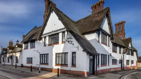 Archivos históricos de Inglaterra Un gran edificio con techo de paja blanco se encuentra en una esquina y tiene un pavimento que rodea el frente del edificio. Hay repisas de ladrillo rojo en las ventanas y tres grandes chimeneas. 