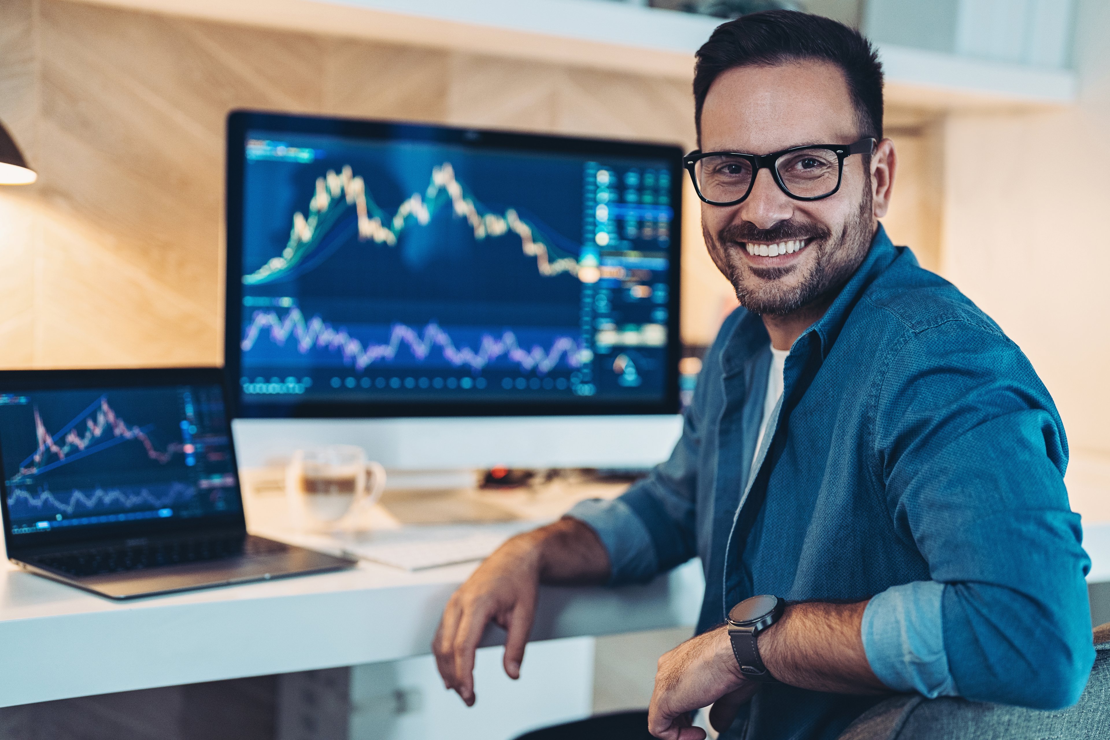 Un inversionista sonriente se sienta frente a dos pantallas de computadora que muestran gráficos de acciones o criptomonedas.