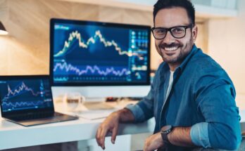 A smiling investor sitting in front of two computer screens, which are displaying stock or cryptocurrency charts.