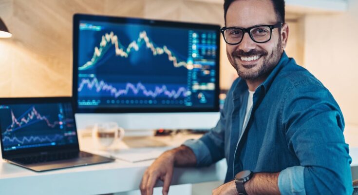 A smiling investor sitting in front of two computer screens, which are displaying stock or cryptocurrency charts.